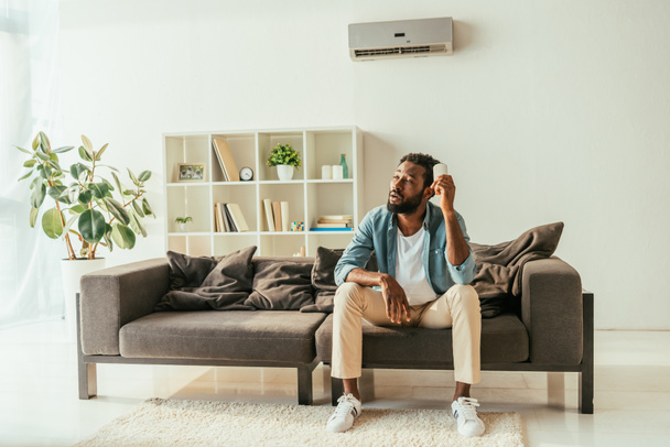 κουρασμένος Αφρικανός Αμερικανός άνθρωπος κάθεται στον καναπέ και κρατώντας κλιματιστικό τηλεχειριστήριο, ενώ υποφέρουν από τη θερμότητα στο σπίτι - Φωτογραφία, εικόνα