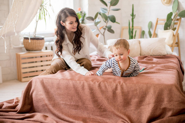 Beautiful young mother with long dark hair in homely comfortable clothes plays with her young son in a cozy bedroom with live green plants and furniture made from natural materials - Foto, Imagem