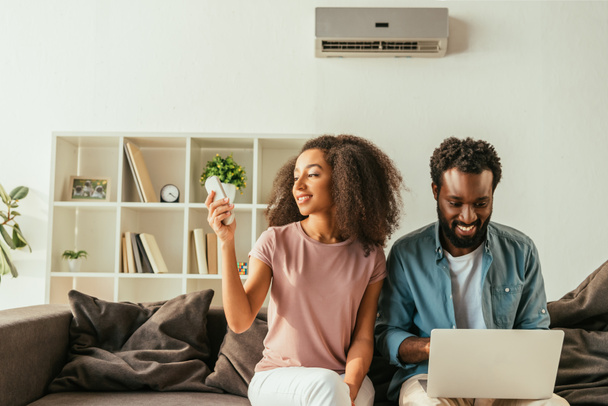 smiling african american man using laptop while sitting on sofa near cheerful african american woman holding air conditioner remote controller - Photo, Image