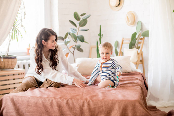 Beautiful young mother with long dark hair in homely comfortable clothes plays with her young son in a cozy bedroom with live green plants and furniture made from natural materials - Foto, Imagen