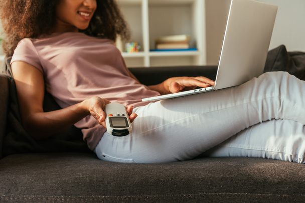 cropped view of smiling african american woman holding air conditioner remote controller while lying on sofa and using laptop - Fotografie, Obrázek
