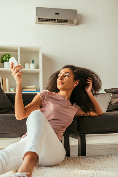 femme afro-américaine insatisfaite assis sur le sol à la maison et en utilisant la télécommande climatiseur
 - Photo, image