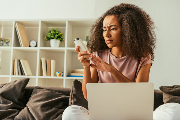 displeased african american woman using air conditioner remote controller while sitting on sofa with laptop - Foto, Imagem