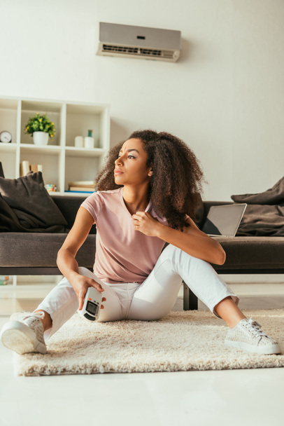 mujer afroamericana joven mirando hacia otro lado y sosteniendo el controlador remoto del aire acondicionado mientras está sentada en el sofá en casa
 - Foto, Imagen