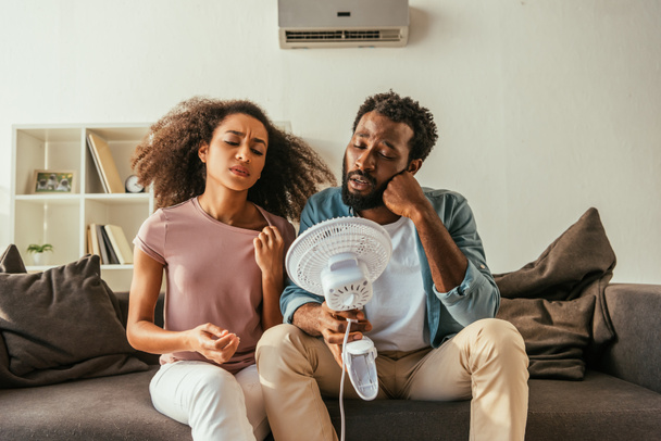 nešťastný afroamerický muž a žena držící elektrický přístroj v době trpící letním teplem doma - Fotografie, Obrázek