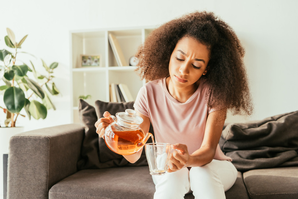 young african american woman pouring tea into cup while sitting on sofa and suffering from heat at home   - Foto, Bild