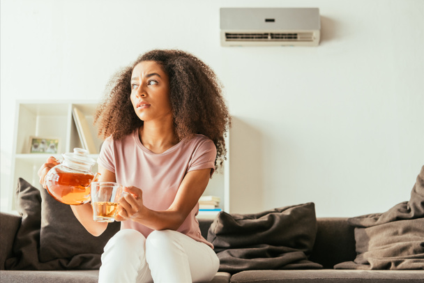 unhappy african american woman pouring tea into cup and looking away while sitting on couch and suffering from summer heat - Photo, Image