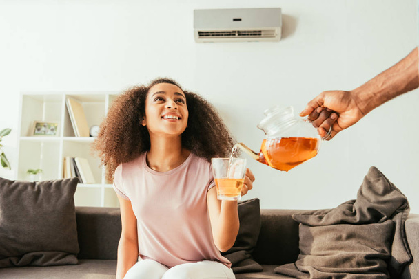 vista recortada del hombre afroamericano vertiendo té en la taza en la mano de la sonriente mujer afroamericana
 - Foto, imagen