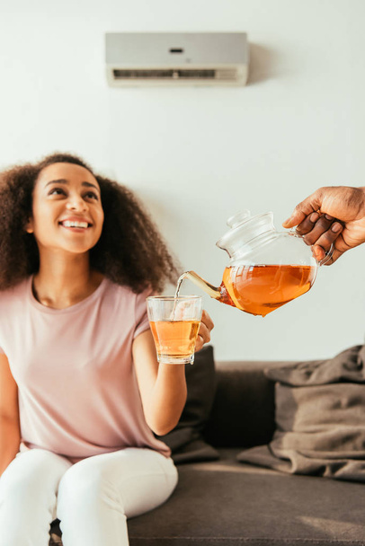 陽気なアフリカ系アメリカ人女性の手にカップにお茶を注ぐアフリカ系アメリカ人男性のトリミングビュー - 写真・画像