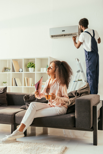 attractive african american woman sitting on sofa with cup of tea, while african american handyman repairing air conditioner - Photo, image