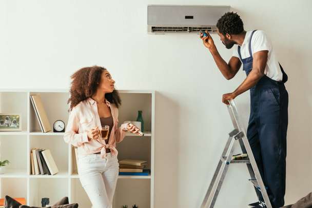 Riparatore afro-americano che ripara il condizionatore d'aria vicino a una donna afro-americana con telecomando
 - Foto, immagini