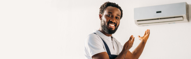 панорамний знімок щасливого афроамериканського ремонтанта, посміхаючись на камеру і вказуючи руками на кондиціонер
 - Фото, зображення
