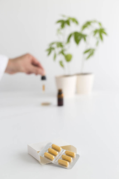 Белый стол с растением марихуаны в керамике. Масло CBD с пипетки в бокал для часов
. - Фото, изображение