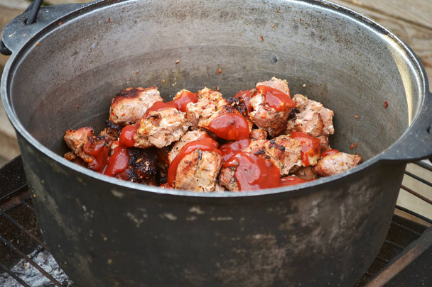 Fleisch vom Grill mit Tomatensauce in einem Topf auf dem Grill, gegen einen Holzzaun - Foto, Bild
