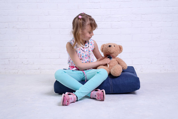 Little girl playing doctor and listening teddy bear with stethoscope against a white brick wall. - Photo, Image