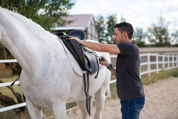 Sivunäkymä nuori kaveri laittaa musta satula takana valkoinen hevonen seisoessaan paddock tilalla
 - Valokuva, kuva