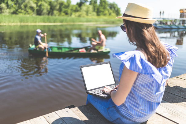 Fashionable woman using laptop while sitting on dock. People in boat and beautiful nature in the background. - Zdjęcie, obraz