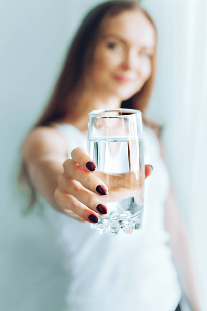 Happy beautiful young woman drinking water. Smiling caucasian female model holding transparent glass in her hand. Closeup. Focus on the arm - Photo, image
