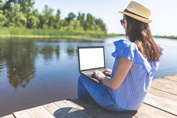 Side view of young woman sitting on pier and using blank screen laptop. Nature and technology concepts. - Фото, изображение