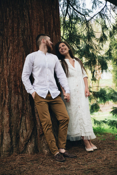 Cheerful young married couple holding hands and  walking in the wood. Sweet pair posing near tree. Beautiful girl and handsome man spending warm day in the forest. Weekend holiday concept - Фото, зображення