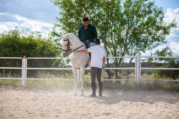 Νεαρός μαθαίνει στους μαύρους να ιππεύουν άλογο στο Paddock τη συννεφιασμένη μέρα στο ράντσο - Φωτογραφία, εικόνα