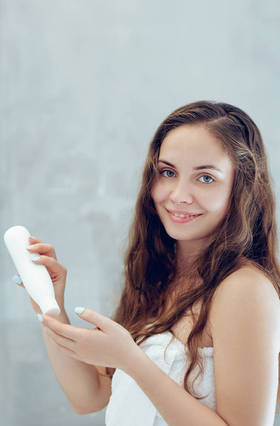 Vlasy. krásná mladá žena, která při stání před zrcadlem v koupelně používá mléko pro vlasy a usmívá se. Péče o vlasy a pokožku. Dívka používá ochranu zvlhčující krém. - Fotografie, Obrázek