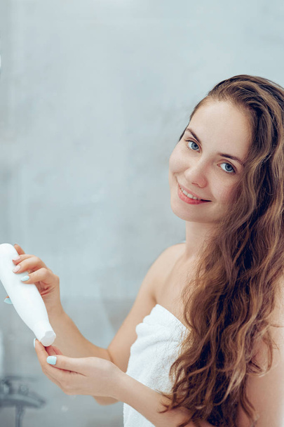 Beautiful young woman applying lotion for hair and smiling while standing in front of the mirror  in bathroom. Care hair and skin.  Girl uses protection moisturizing cream. - Fotografie, Obrázek