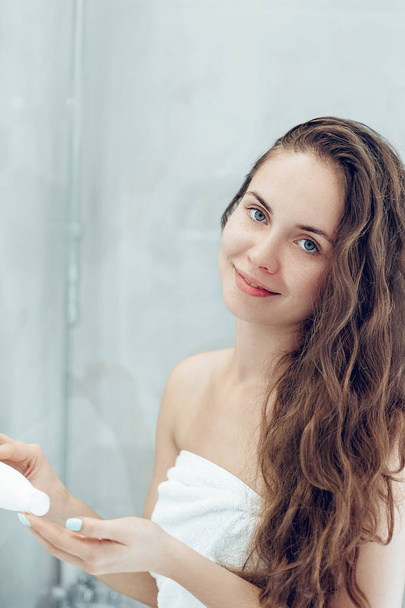 Beautiful young woman applying lotion for hair and smiling while standing in front of the mirror  in bathroom. Care hair and skin.  Girl uses protection moisturizing cream. - Φωτογραφία, εικόνα