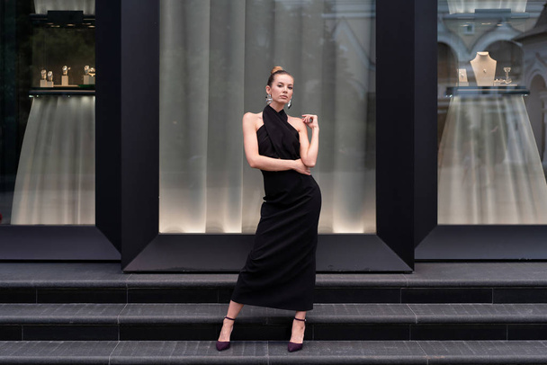 Beautiful girl in a long black dress posing in full growth near the boutique. Graceful interesting poses. - Fotoğraf, Görsel