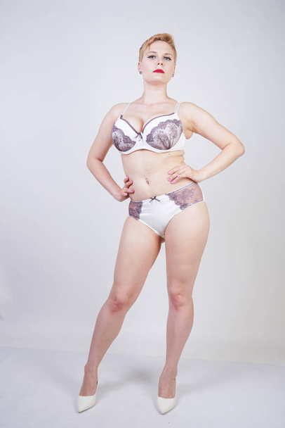 charming young woman with short blonde hair and curvy body posing in white lingerie with purple lace on the solid background in the Studio. plus size adult girl wearing underwear and standing alone. - Photo, Image