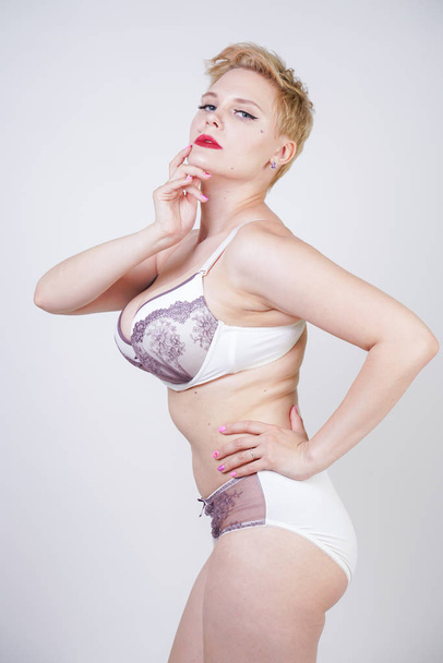 charming young woman with short blonde hair and curvy body posing in white lingerie with purple lace on the solid background in the Studio. plus size adult girl wearing underwear and standing alone. - Фото, изображение