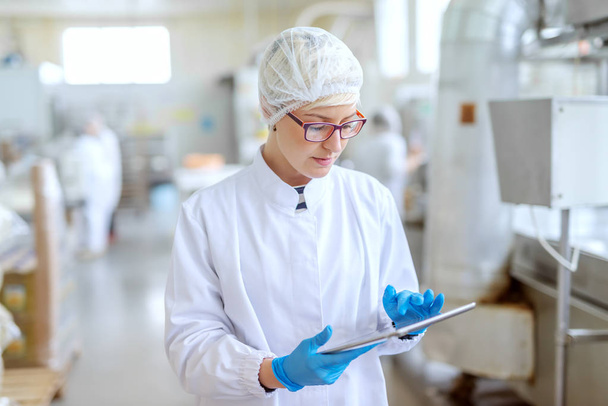 Superviseur en uniforme stérile et avec lunettes utilisant une tablette pour contrôler le flux de travail dans l'usine alimentaire
. - Photo, image