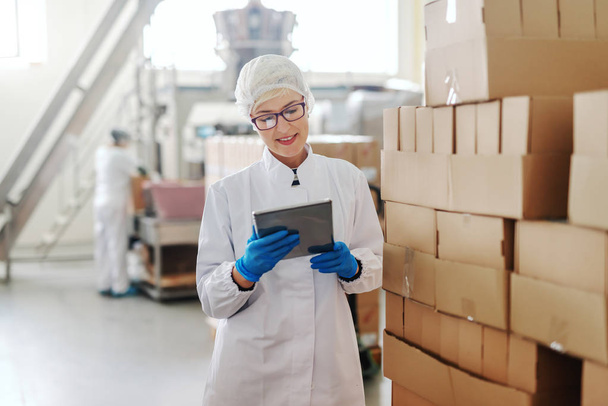 Kaukasische blonde werknemer in steriele uniform permanent en het omgaan met de logistiek van producten. Voedsel fabriek interieur. In hand tablet. - Foto, afbeelding