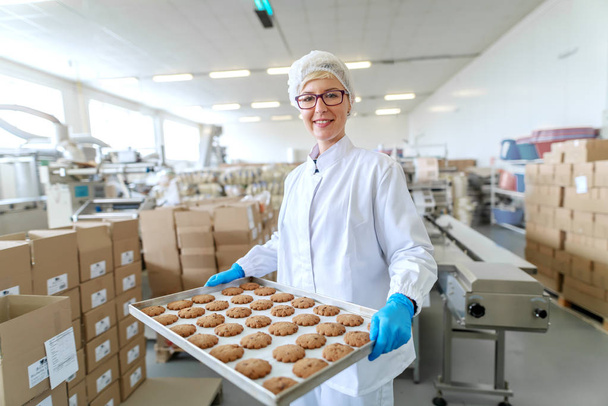 Usměvavý blonďatý zaměstnanec ve sterilní uniformě a s brýlemi stojící a držící podnos s koláči. Interiér potravinářského podniku. Na pozadí ostatní zaměstnanci pracující. - Fotografie, Obrázek