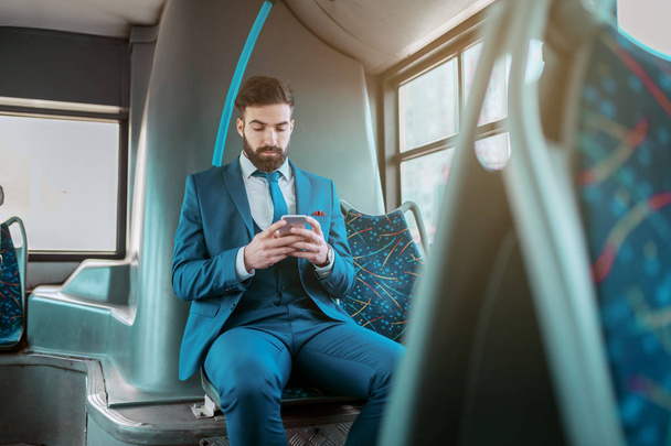 Young attractive serious Caucasian bearded businessman in blue suit sitting in public bus and using smart phone. - Photo, Image