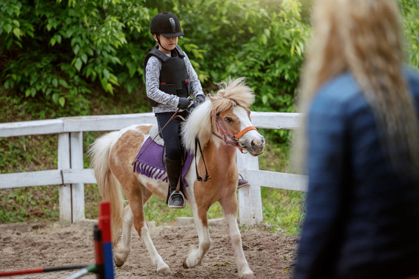 Fille caucasienne avec casque et gilet de protection sur l'équitation cheval poney blanc et brun mignon. Journée ensoleillée sur le concept du ranch. - Photo, image