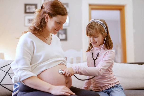 Beautiful Caucasian pregnant mother sitting in living room with her daughter. Daughter listening with stethoscope baby's heartbeats. Living room interior. - 写真・画像