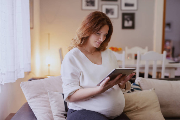 Beautiful Caucasian pregnant woman sitting in living room in sofa and using tablet. - Foto, imagen