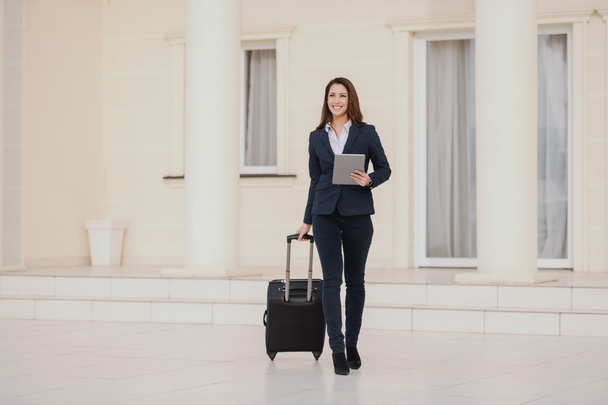 Young smiling brunette dressed smart casual walking, holding luggage and tablet. Business trip concept. - 写真・画像