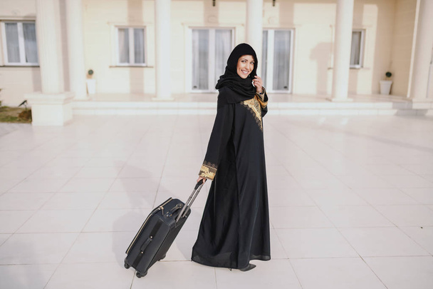 Hymyilevä musliminainen perinteisessä kulumisessa seisoo talonsa edessä ja pitää matkatavarat
. - Valokuva, kuva