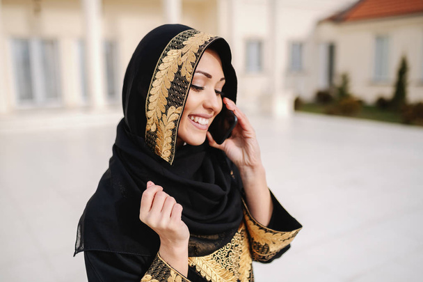 Портрет очаровательной улыбающейся мусульманки, одетой в традиционную одежду, с помощью смартфона, стоя на улице
. - Фото, изображение