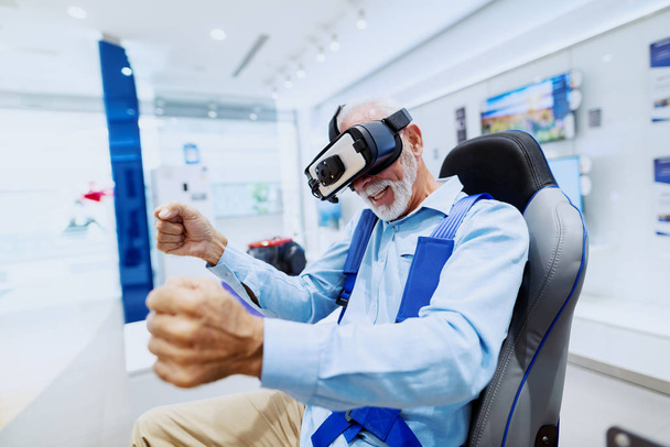 Захоплений бородатий старший чоловік сидить у кріслі і випробовує окуляри віртуальної реальності. Інтер'єр магазину
. - Фото, зображення