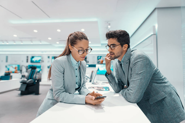 Carino coppia multiculturale in abbigliamento formale discutendo di smart phone che vogliono comprare. Tech negozio interno
. - Foto, immagini