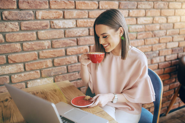 Smiling Caucasian brunette dressed elegant drinking coffee in cafeteria. On desk laptop. - Fotografie, Obrázek