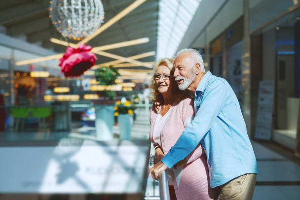Senior paar in liefde flirten terwijl leunend op reling in winkelcentrum. - Foto, afbeelding