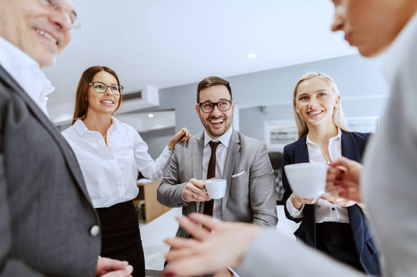 Grupa uśmiechniętych radosnych kolegów siedzących i stojących w miejscu pracy, rozmawiając i pijąc kawę na pauzy. - Zdjęcie, obraz