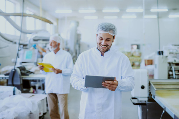 Young Caucasian smiling supervisor in sterile white uniform using tablet while standing in food plant. In background older one worker controlling machine. - Photo, Image