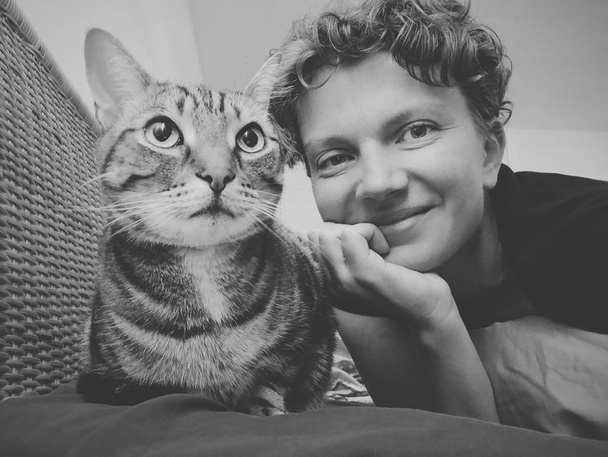 Black and white selfie portrait of cat and its owner girl lying on couch sofa looking in camera. Smiling Caucasian woman with kitten pet feline animal. Authentic style real person lifestyle. - Photo, Image