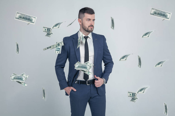 πορτρέτο του κομψού επιχειρηματία με πτώση δολαρίου τραπεζογραμμάτια απομονώνονται σε γκρι φόντο. ντυμένη με ένα κοστούμι με ένα κομψό χτένισμα και ένα μούσι - Φωτογραφία, εικόνα