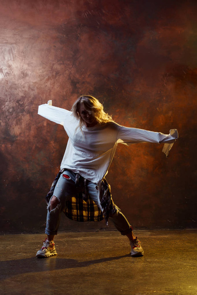 Photo of dancing long-haired blonde girl in torn blue jeans on brown background - Zdjęcie, obraz